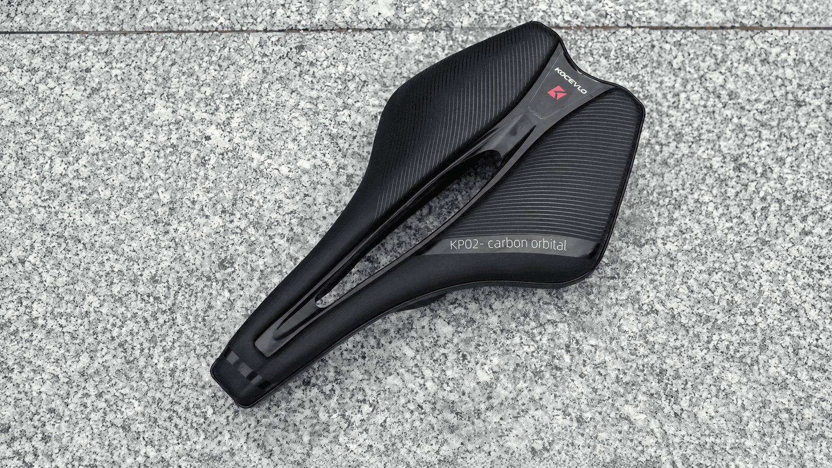Top view of KOCEVLO ultralight carbon road bike saddle on grey stone pavement, hollow cutout ergonomic bicycle seat with brand logo